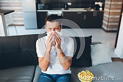Young man watch tv in his own apartment. Guy sneezing into white napkin. Sick and ill at home. Suffer from headache or Stock Photo