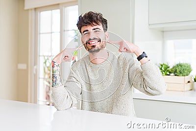 Young man wearing casual sweater sitting on white table smiling confident showing and pointing with fingers teeth and mouth Stock Photo