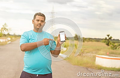 Young man wearing jogging dress showing smartphone screen by pointing with hand and finger at park - concept of recommended Stock Photo