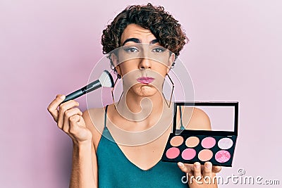 Young man wearing woman make up holding makeup brush and blush puffing cheeks with funny face Stock Photo