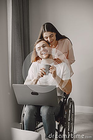 Young man in wheelchair using laptop and girlfriend hugging him at home Stock Photo
