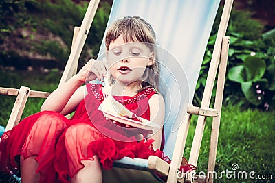 Young mannered girl in red dress relaxing in chaise lounge eat cake during outdoor party in the garden Stock Photo