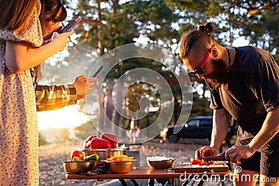 A young man with a beard prepares a salad of vegetables in nature, the girl takes pictures on the phone. Rest friends in the Stock Photo