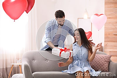 Young man presenting gift to his girlfriend in room decorated with heart shaped balloons. Valentine`s day celebration Stock Photo