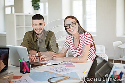 Young man and woman in striped t-shirt and eyeglasses happily looking in camera while working together. Group of cool Stock Photo