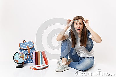 Young mistrustful puzzled woman student in denim clothes removing glasses sitting near globe, backpack, school books Stock Photo