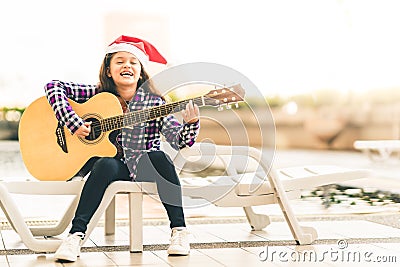 Young mixed race girl playing guitar, singing and smiling joyfully by swimming pool, with christmas santa hat Stock Photo