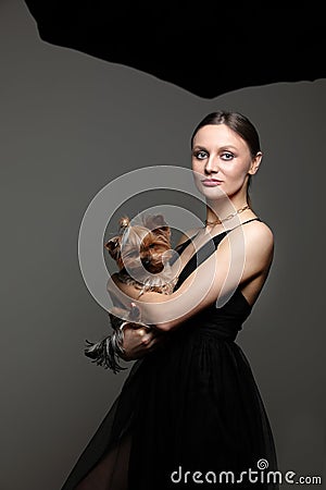 Young model woman with make up, hairstyle wearing in fashionable blak dress keeps a Yorkshire terrier on her hands over Stock Photo