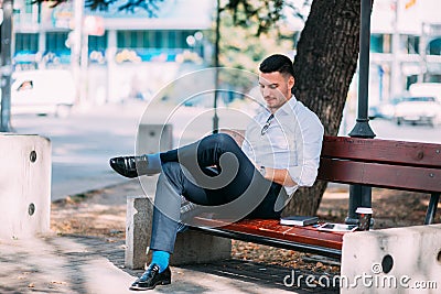 Young modern businessman is sitting in the park on a bench with crossed legs and he is looking curious and serious Stock Photo