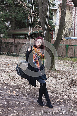 Young modern caucasian woman in black coat and colorful scarf outdoor pose on swing in park Stock Photo