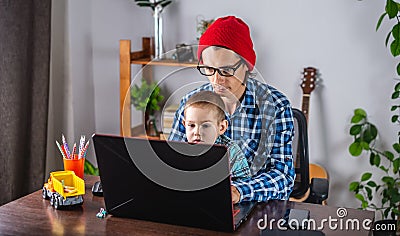 Modern man is working on a laptop, and his little son is sitting on his lap. Concept of family and remote work from home Stock Photo