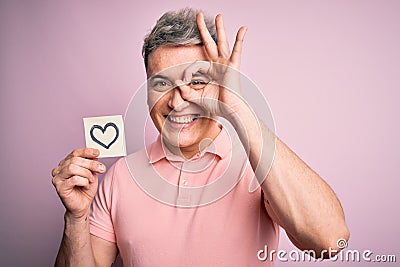 Young modern romantic man holding heart shape drawing over isolated pink background with happy face smiling doing ok sign with Stock Photo