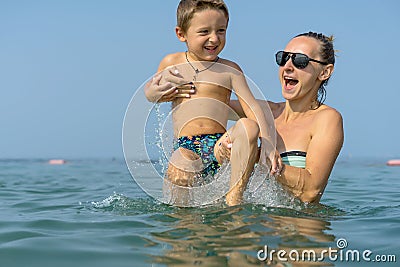 Young mother in black sunglasses and smiling baby boy son in green baseball cap playing in the sea in the day time. Positive human Stock Photo