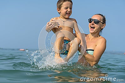 Young mother in black sunglasses and smiling baby boy son in green baseball cap playing in the sea in the day time. Positive human Stock Photo