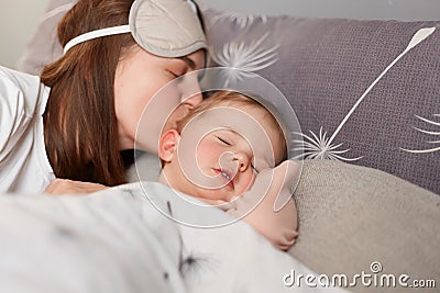 Young mother dark haired woman kissing sweet little baby, cute kid laying near mom and sleeping while parent kissing her, home Stock Photo