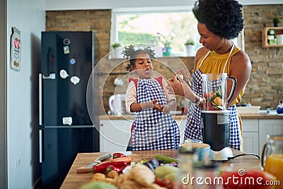 Young mother in the home kitchen with her little daughter, cutting up vegetables and tasting cucumber slices, girl acting silly Stock Photo