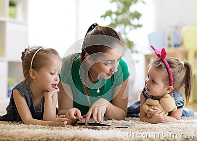 Young mother reading a book to her kids daughters. Children and mom lying on rug in sunny living room. Stock Photo