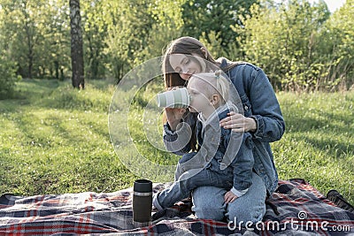 Young mother and small daughter on picnic. Girl drinks from cup. Green park on sunny spring day. Little girl sits on her mother`s Stock Photo