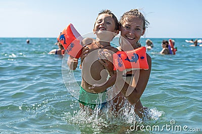Young mother and smiling baby boy son in green baseball cap playing in the sea in the day time. Positive human emotions, feelings, Stock Photo