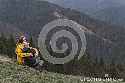 Young mother with son sit on hillside. Mother and son rest and sit in an embrace on mountains background Stock Photo