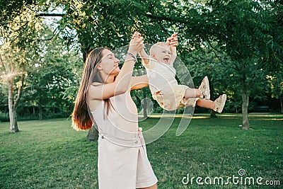 Young mother spinning toddler baby boy outdoors. Parent playing with child son in park. Authentic lifestyle funny moment. Happy Stock Photo