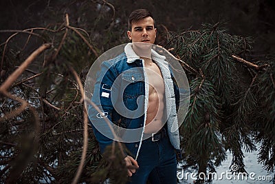 Young muscular man in unbuttoned jacket with bared breast stands next to pine tree in winter forest. Stock Photo