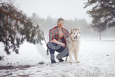 Young muscular man in unbuttoned shirt sits and hugs dog Malamute at walk in winter misty forest. Stock Photo