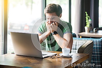 Young nervous businessman in green t-shirt sitting and working on laptop, bitting his nails and looking at screen with worry face Stock Photo