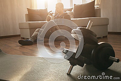 Young ordinary man go in for sport at home. Tired workout freshman sit on floor alone and has rest after exercise. Drink Stock Photo