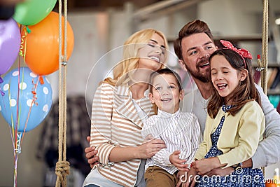 Young parents and their kids enjoy posing for a photo while sitting on the swing together at home. Family, home, playtime Stock Photo