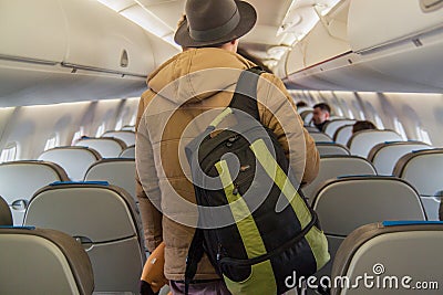 Handsome young passenger in a warm jacket and hat stands with a backpack in the cabin of airplane. Stock Photo