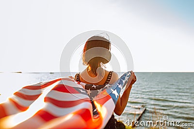 Young patriotic woman holds American flag in the wind on the beach on a sunset wearing medical protictive mask. 4th July, Stock Photo