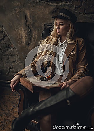 Young pensive woman in image of Sherlock Holmes sits in armchair and holds photoalbum in her hands with magnifier on background of Stock Photo