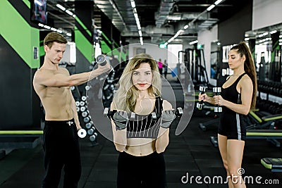 Young people pumping up muscles with dumbbells. Pretty blond fitness girl exercising with barbell in gym together with Stock Photo