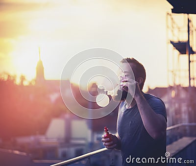 Young people spend time on the terrace of the house, blowing bubbles with the help of vaporizer against backdrop of the sunset Stock Photo