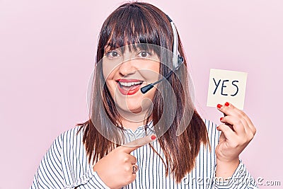 Young plus size woman wearing call center agent headset holding yes reminder smiling happy pointing with hand and finger Stock Photo