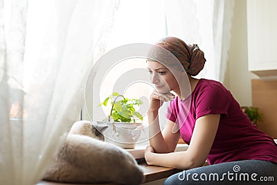 Young positive adult female cancer patient sitting in the kitchen by a window with her pet cat. Stock Photo