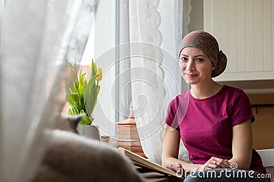 Young positive adult female cancer patient sitting in the kitchen by a window reading a book, smiling. Stock Photo