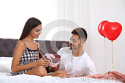 Young presenting gift to his girlfriend in bedroom decorated with air balloons. Celebration of Saint Valentine`s Day Stock Photo