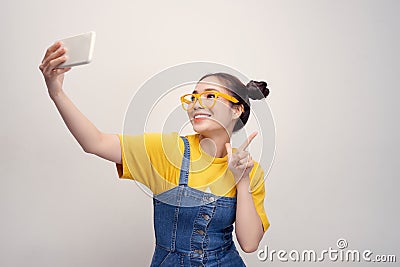 Young pretty Asian girl who wearing a jeans dungaree and yellow glasses, taking selfie, smiling Stock Photo