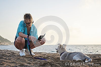 Young pretty blond woman, taking picture on her phone of her small husky puppy. Owner and the light grey dog, playing on the sandy Stock Photo
