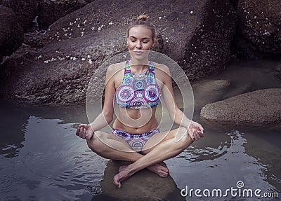 Young pretty blonde female wearing colorful bikini sitting on the rock and doing meditation yoga. Stock Photo