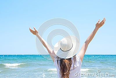 Young Pretty Caucasian Woman with Long Chestnut Hair in Hat Hands Lifted up in the Air Stands at Beach Looks at Turquoise Sea Stock Photo