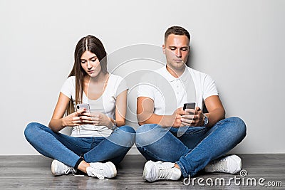 Young pretty couple typing on phone while sitting on the floor isolated on gray background Stock Photo