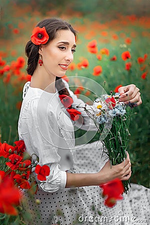 Young pretty girl Slavic or Ukrainian posing in folk dress on a flowering poppy field. Female holding a bouquet of Stock Photo