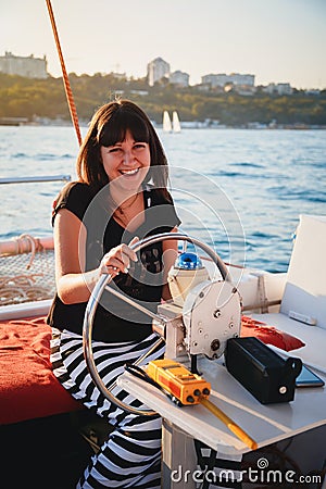 Young pretty smiling woman in black shirt and striped skirt driving luxury yacht in sea, sunset Stock Photo