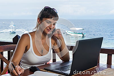 Young pretty woman freelancer writer working with laptop notepad and phone in front of blue tropical sea Stock Photo