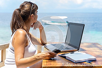 Young pretty woman freelancer writer working with laptop notepad and phone infront of blue tropical sea Stock Photo