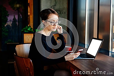 Young pretty woman in glasses works on a laptop, uses a smartphone, a freelancer, a computer, financial analyst, a sales manager. Stock Photo