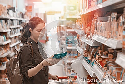 A young pretty woman holds a grocery cart and carefully chooses the products in the store. Light. Close up Stock Photo